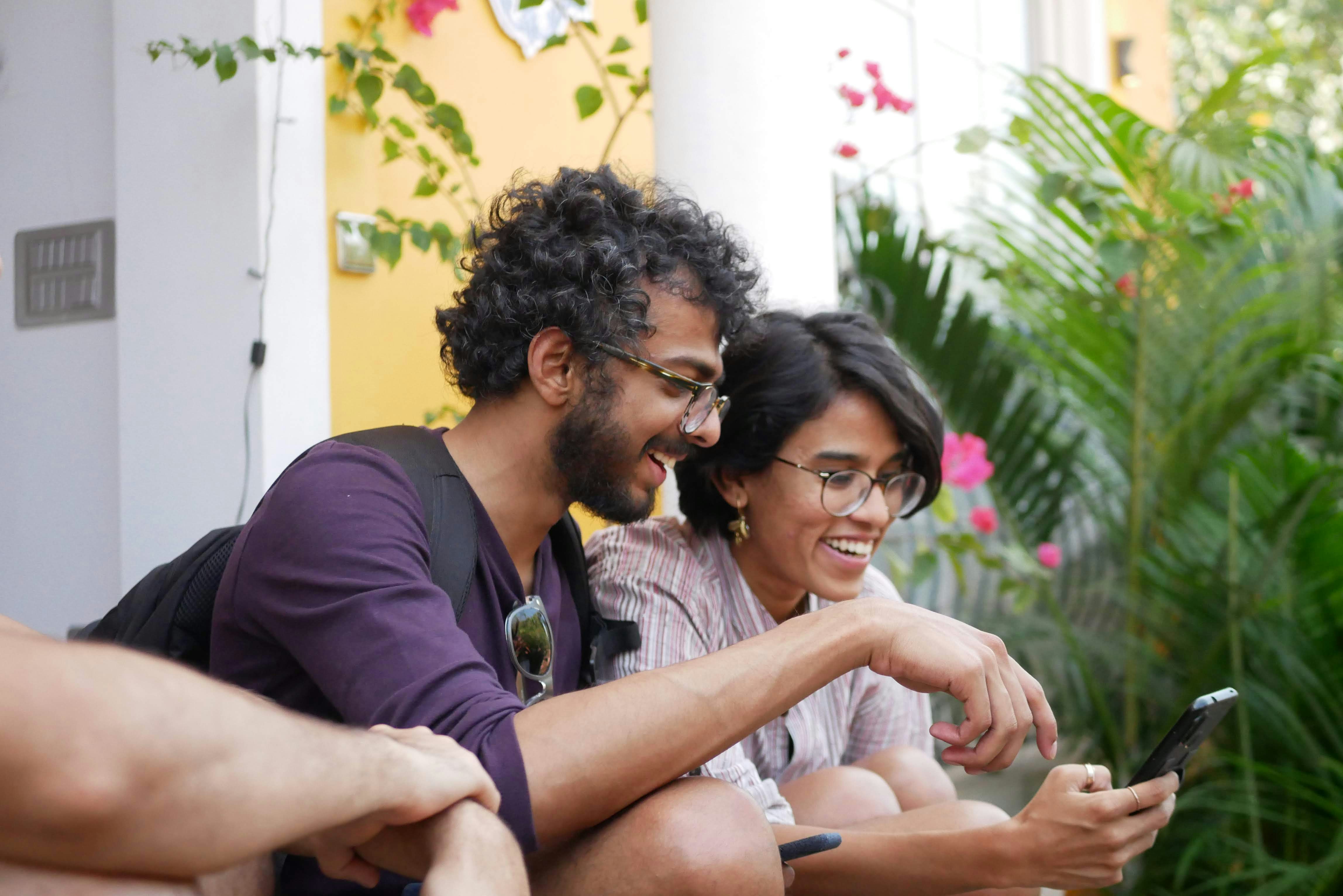 Two people laughing together while looking at a phone, outdoors among flowers